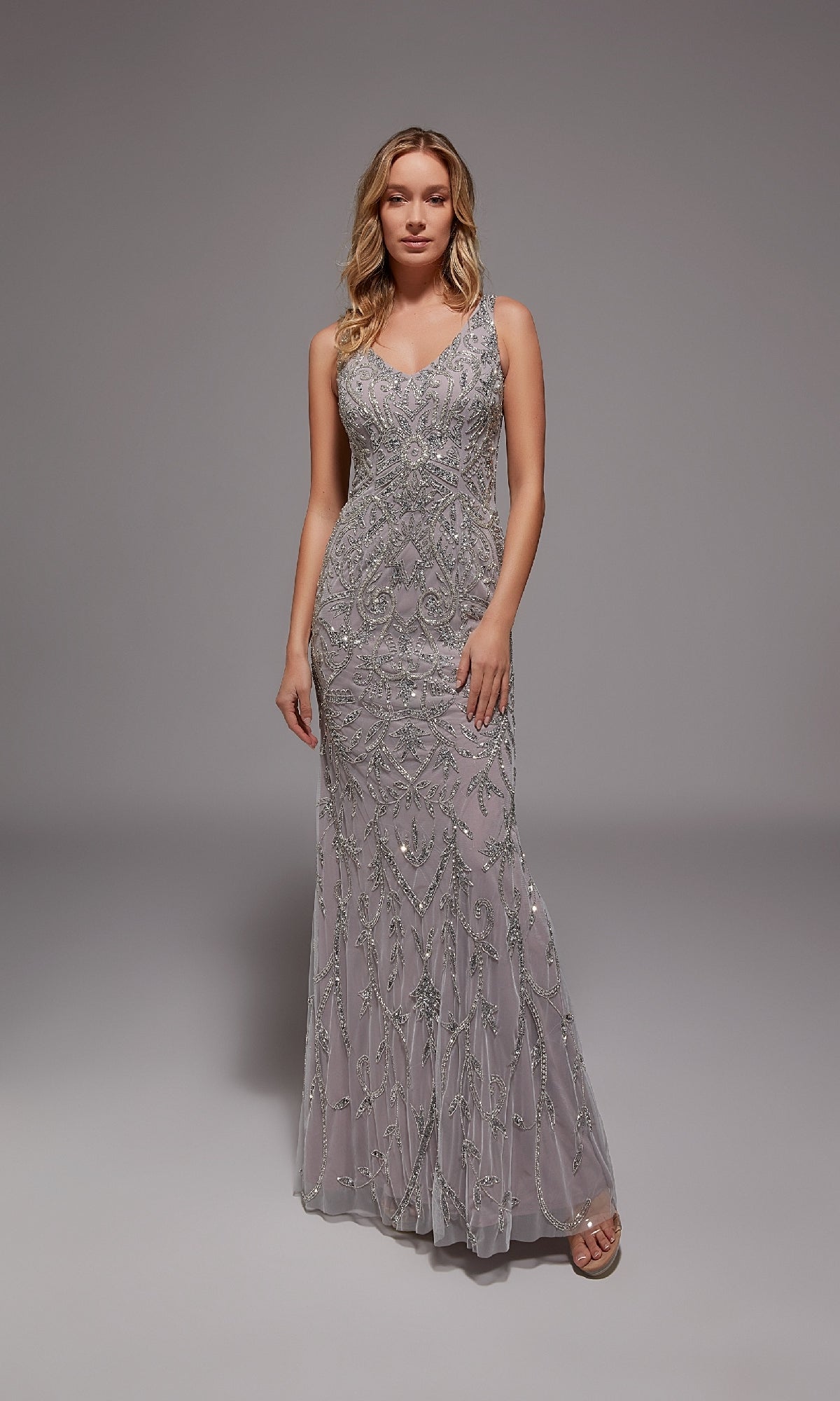 A woman stands in a studio wearing the Long Formal Dress: Alyce 27799—a floor-length, silver/lilac evening gown with beading, fitted silhouette, and V-neckline.